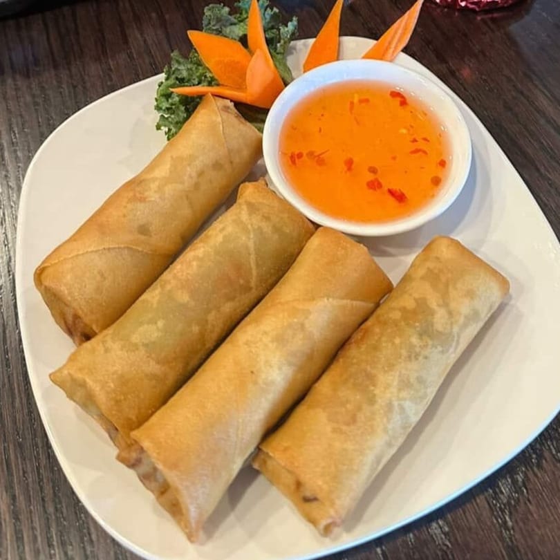 Four golden-brown spring rolls with a side of orange sweet chili sauce and garnish on a white plate.